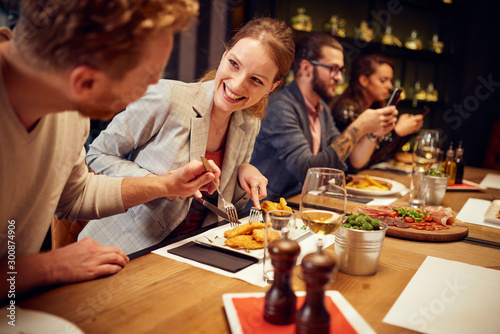 Handsome caucasian ginger taking food out of his girlfriend's plate while sitting in restaurant for dinner. In background are their friends.