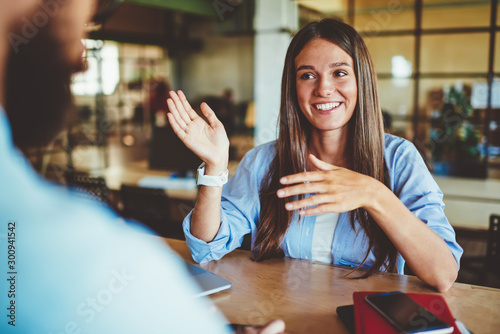 Happy successful hipster girl explaining information to male friend sitting in front, positive man and woman talking and discussing ideas for new startup project at spending time in coworking space