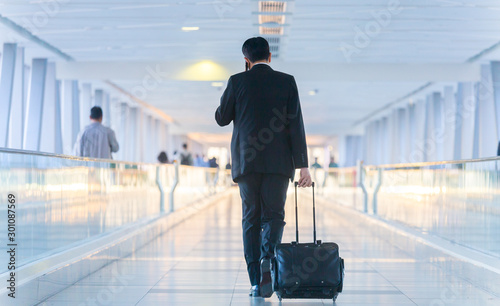 Rear view of unrecognizable formaly dressed businessman walking and wheeling a trolley suitcase at the lobby, talking on a mobile phone. Business travel concept.