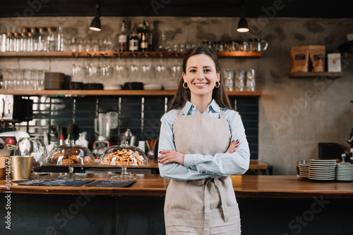 smiling barista in apron standing with crossed arms near bar counter