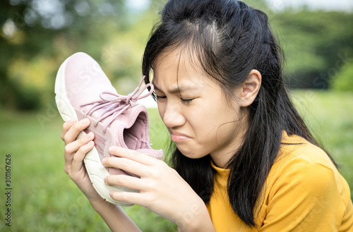 Unhappy asian female teenage is sniffing her sneakers,hold stinky shoe in her hand with disgust,unpleasant smell because of hot weather or after exercise,sad child girl with bad smell,accumulated dirt