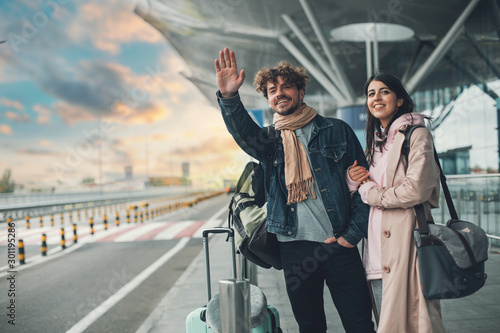 Young man and woman stand outside airport and wait for taxi cab. Guy wave with hand. After vacation or travel. Georgian woman hold his hand and smile. Sunset outside. Evening.