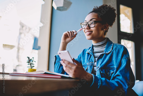 Positive contemplative hipster girl in stylish eyewear for vision correction looking away and thinking on idea for content to web page during break from exam preparation with knowledge textbook