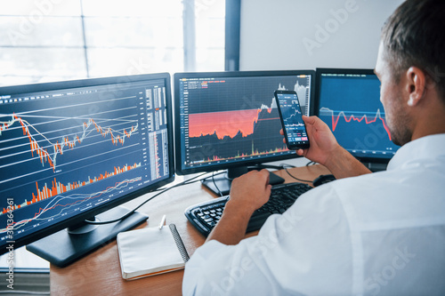 Young businessman in formal clothes is in office with multiple screens. Conception of exchange and money