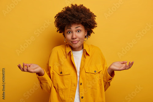 Puzzled clueless African American lady shrugs shoulders and expresses uncertainty, makes decision, wears fashionable clothing, spreads palms sideways isolated on yellow wall. Life perception, attitude