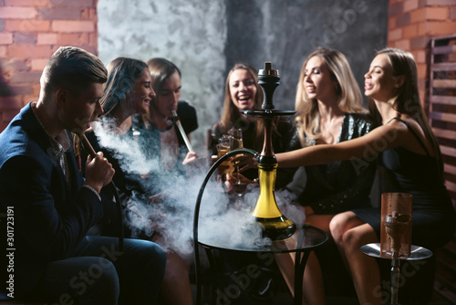 Friends party in hookah lounge. Group of people women and men smoking shisha in cafe or bar, making smoke clouds, having fun, smiling. Relax concept. Friendship