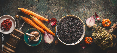Black lentils with cooking ingredients, vegetables, herbs and spices for tasty vegan meals on dark background. Top view. Healthy vegetarian eating concept. Horizontal banner. Plant based protein