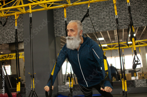 Bearded senior sportsman training with trx resistance bands in gym.