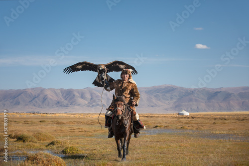 A proud young kazakh eagle hunter posing with his golden eagle on horseback on the backdrop of blue sky. Ulgii, Mongolia.