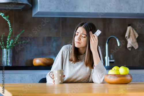 Brunette girl is sitting at table in kitchen and holding hand on head temple. Young woman is feeling bad and going to take painkiller pills. Sudden attack of migraine and headache. Effects of stress.