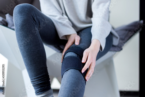 Closeup female leg in grey leggings dressed in knee brace to help promote recovery of bones, muscles, ligaments. Woman is feeling pain in joints after injury. Arthritis and meniscus diseases concept.