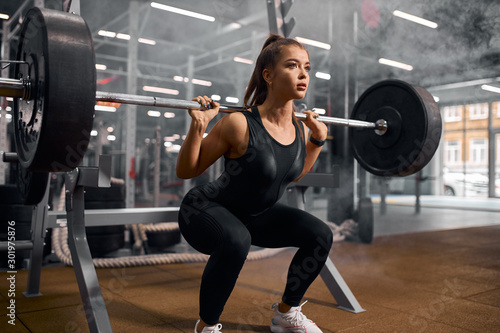 Charming strong female powerlifter dressed in black sportswear and white sneakers, doing squats, trying to stand with heavy barbell, professional sport concept, indoor shot
