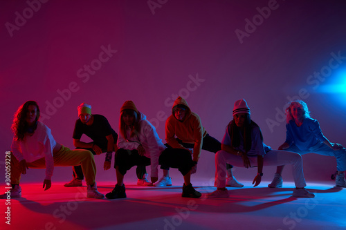 Big group of young people sitting on squat on dark lighted stage, waiting for beginning of music, ready to start dancing, hip hop, street dance, break dancers