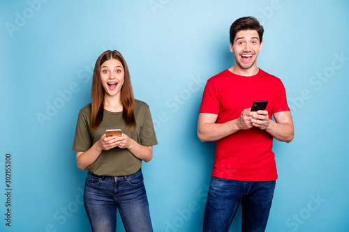 Portrait of amazed crazy two married people use smart phone read social network newas impressed got like incredible feedback wear green t-shirt denim jeans isolated blue color background