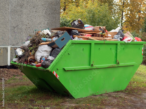 Big green color metal skip container full of tras by a house building.