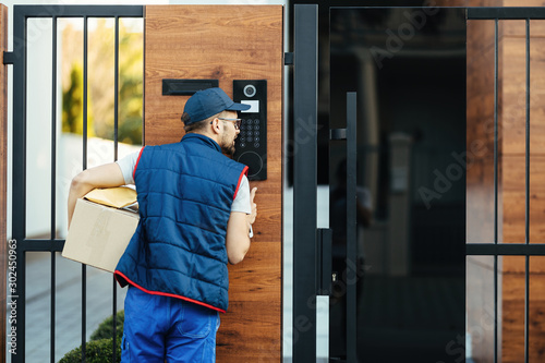 Rear view of a courier ringing on customer's intercom while making home delivery.