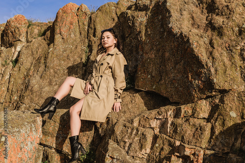 young beautiful brunette in a beige spring coat in the field under the autumn sun. attractive young woman in autumn image on a walk posing sexy against the background of rock and desert