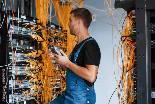 Young man in uniform with measuring device works with internet equipment and wires in server room