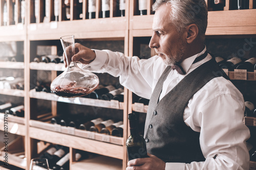 Sommelier Concept. Senior man standing looking at wine decanter checking sediment concentrated side view