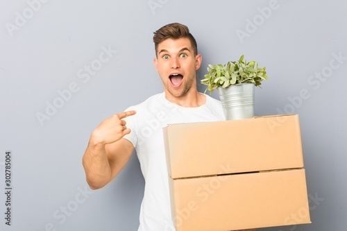 Young man doing a move holding boxes surprised pointing at himself, smiling broadly.