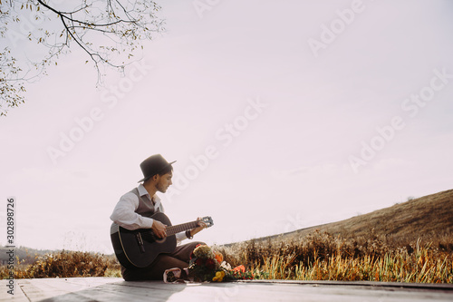 Young man playing guitar sitting on the bank of a mountain river on a background of rocks and forest. Handsome hippie style guitarist engrossed on music outdoors. Concept of freedom relaxation. Place