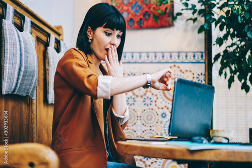 Shocked woman checking time on wearable wristwatch after working on laptop device at bohemian cafeteria, amazed caucasian woman wonder of deadline spending time for distance job at cozy cafe