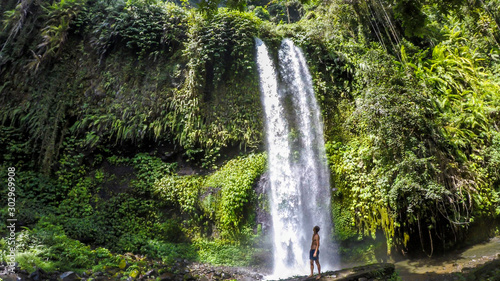 A man in swimsuit standing under two levelled waterfall in Lombok, Indonesia. Tiu Kelep Waterfall is surrounded by lush green plants from each side. Long and powerful waterfall. Beauty of the nature.