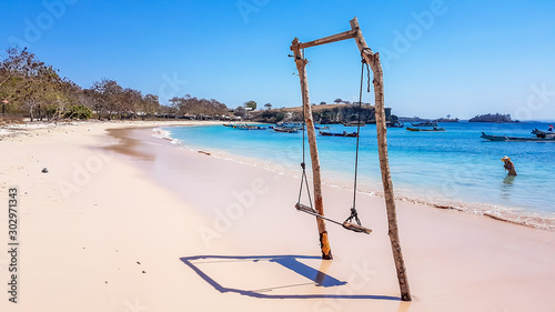 A swing placed on the seashore of Pink Beach, Lombok, Indonesia. The swing has very simple wood construction. Waves gently wash the pillars of it. In the back there are few boats anchored in the bay.