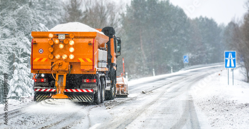 Snow plow on highway salting road. Orange truck deicing street at snowing time. Crystals dropping on asphalt. Maintenance winter vehicle in action.