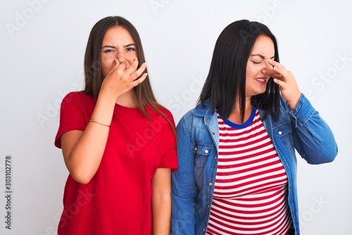 Young beautiful women wearing casual clothes standing over isolated white background smelling something stinky and disgusting, intolerable smell, holding breath with fingers on nose. Bad smells 