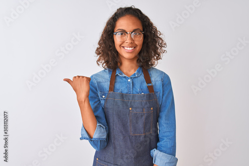 Young brazilian chef woman wearing apron and glasses over isolated white background smiling with happy face looking and pointing to the side with thumb up.