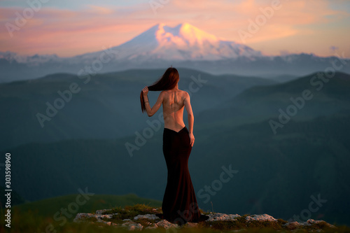 A beautiful young girl with a naked back is standing in an evening dress in the mountains of the Caucasus while admiring the sunset. girl looks at a sleeping volcano covered in snow and ice