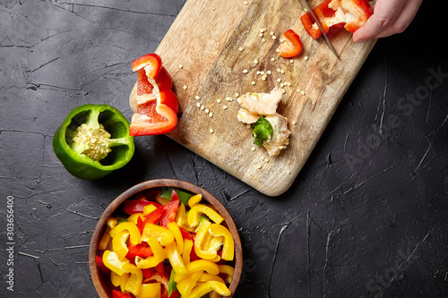 Wooden cutting board and bowl with pieces of different bell peppers on black background, top view. Cooking vegetable salad, healthy food, diet dish. Chopped red, green, yellow sweet peppers