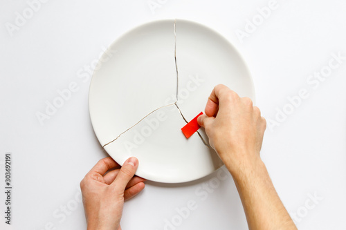 Top view of man hands holding a broken white plate, glues parts with red tape. Metaphor for divorce, relationships, friendships, crack in marriage. Isolated on white background, copy space. 