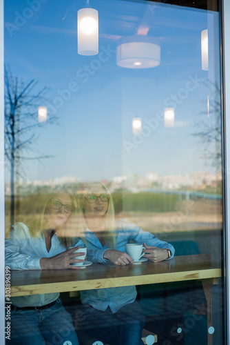 Two friends are enjoying coffee in a coffee shop, looking through a glass with reflections, sitting at a table chatting. Elderly Mother and adult daughter in a cafe. Mothers Day. Family resemblance.