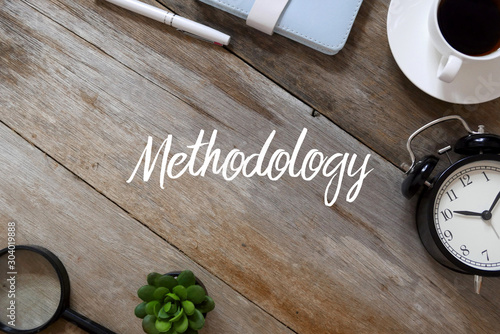 Top view of pen,notebook, a cup of coffee, clock,plant and magnifying glass on wooden background written with Methodology.