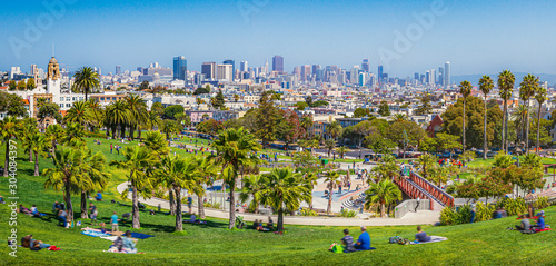 Panoramic view of local people enjoying the sunny summer weather at Mission Dolores Park on a beautiful day with clear blue sky with the skyline of San Francisco in the background, California, USA