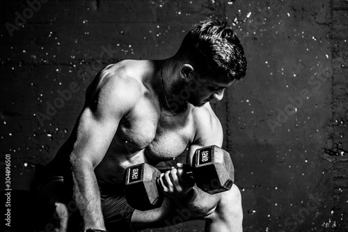 Young strong muscular sweaty fit man biceps muscle cross workout training with heavy dumbbell weight in the gym dark image with shadows real people black and white