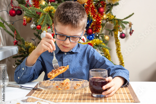 Chłopiec w okularach siedzi przy stole i ze smakiem zjada obiad. Bożonarodzeniowa choinka w tle.