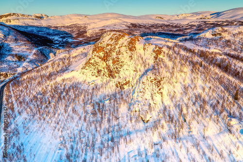AERIAL: Sunset golden light over Norwegian snowy winter mountains with birche and pine trees, early spring, calm blue skies. Rossvatnet lake area, Hattfjelldal municipality, Northern Norway