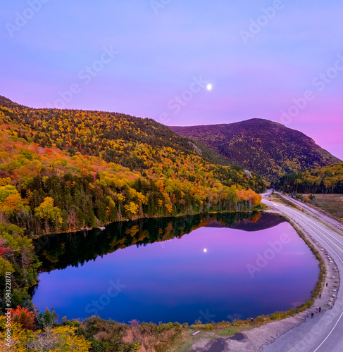 Aerial drone view of fall foliage in mountains with reflection of pink sky and moon in pond