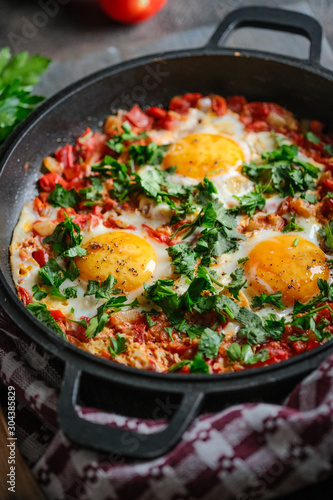 Traditional shakshuka with eggs, tomato, and parsley in a iron pan on a dark background