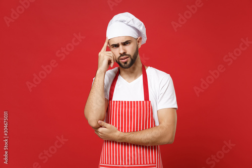Pensive young bearded male chef cook or baker man in striped apron toque chefs hat posing isolated on red background in studio. Cooking food concept. Mock up copy space. Pointing index finger on head.