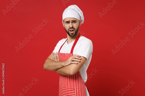 Irritated young bearded male chef cook baker man in striped apron white t-shirt toque chefs hat posing isolated on red wall background. Cooking food concept. Mock up copy space. Holding hands crossed.