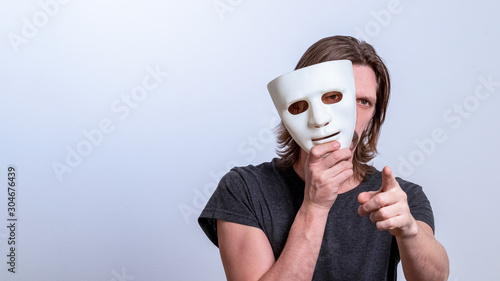 A long-haired man in a gray T-shirt looks out from behind a white mask and points his finger at the camera. The concept is you next, fraud. Copy space. Gender or split personality. Caution Warning.