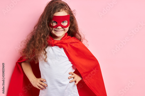 Portrait of confident smiling kid superhero wearing red cloak and mask on eyes imagines herself as hero, stand isolated ove pink background