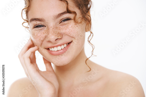 Headshot satisfied tender smiling redhead cute girl freckles curly-haired, standing naked white background laughing happily gently touch cheek look after skin like result apply cosmetology product