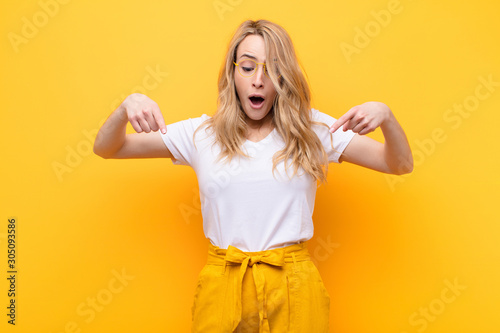 young pretty blonde woman feeling shocked, open-mouthed and amazed, looking and pointing downwards in disbelief and surprise against flat color wall