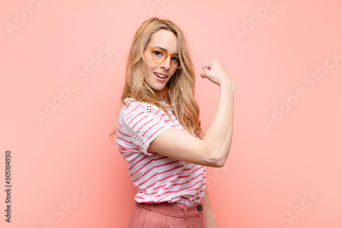 young pretty blonde woman feeling happy, satisfied and powerful, flexing fit and muscular biceps, looking strong after the gym against flat color wall