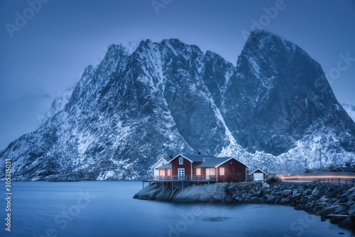 Red rorbu on sea coast and snow covered mountain at dusk. Lofoten islands, Norway. Moody winter landscape with traditional norwegian rorbuer, water, snowy rocks at night. Old fishermen's houses
